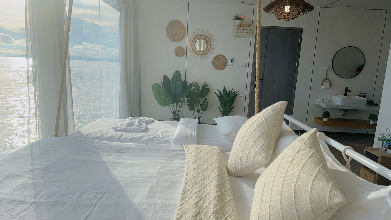 An inviting bed inside a warm bedroom at Sunny Holiday near Bum Bum Island showcasing neatly folded pink towels, blue headboard drapes, rich mahogany style wooden floors, and a/c.