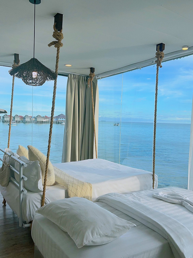 A spacious bedroom at Sunny Holiday over the water, offering a large swinging bed next to a single bed, modern style woven lighting, polished wood floors, A/C, and floor to ceiling glass windows.