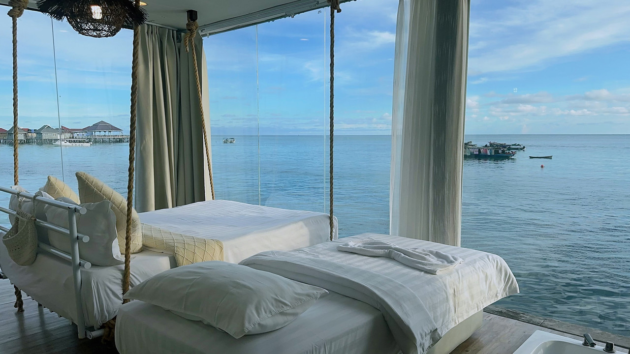 A stunning water resort bedroom at Sunny Holiday in the Celebes Sea, boasting a large swinging bed and a single bed facing panoramic glass windows, polished timber floors, and air conditioning.