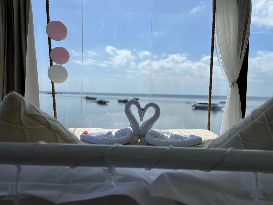 A scenic bedroom setup viewed from behind the bed, showing towel swans against expansive floor-to-ceiling glass doors that reveal a sweeping seaview over Tun Sakaran Marine Park, showcasing modern beachfront style at Sunny Holiday.