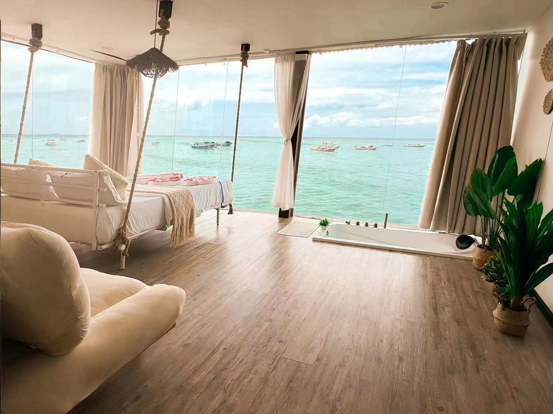 A unique bedroom at Sunny Holiday near Semporna featuring a large swinging bed facing panoramic glass windows, a sunken bathtub, modern style decor, smooth wood floors, and air con.