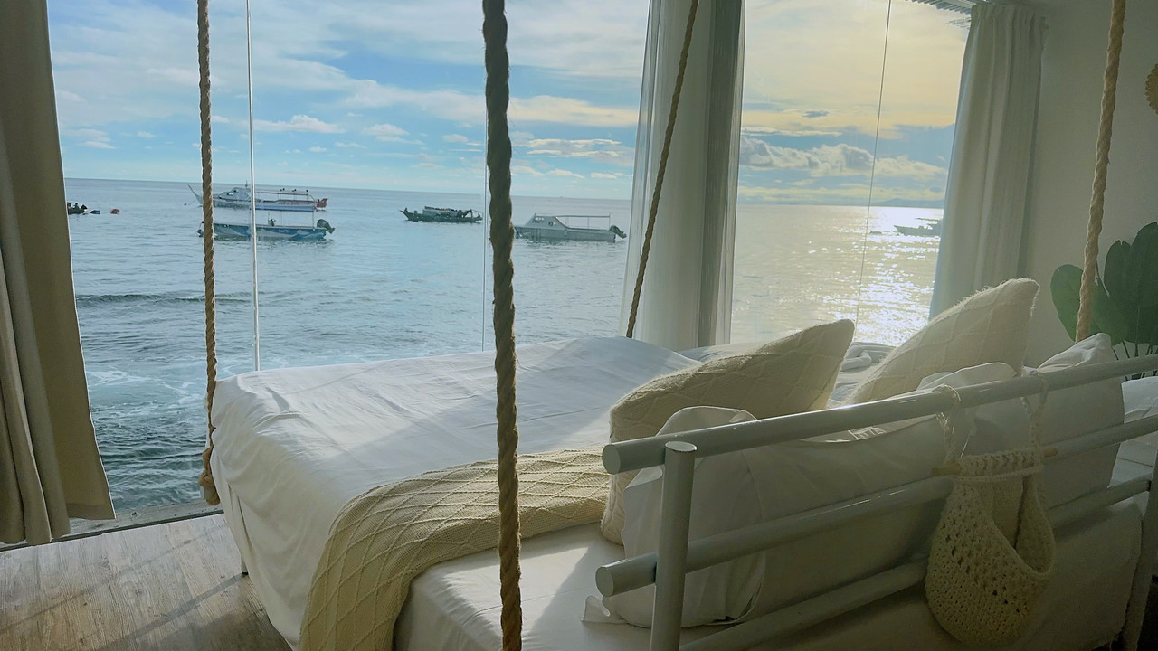 A beautiful bedroom at Sunny Holiday near Tun Sakaran Marine Park, offering a large swinging bed facing panoramic glass windows framing the seaviews, modern style decor, sleek wood floors, and A/C.