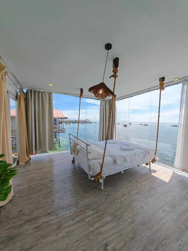 An expansive bedroom at Sunny Holiday near Tun Sakaran Marine Park, highlighting a large swinging bed facing the ocean, traditional style lighting, smooth wood floors, AC, and panoramic windows.